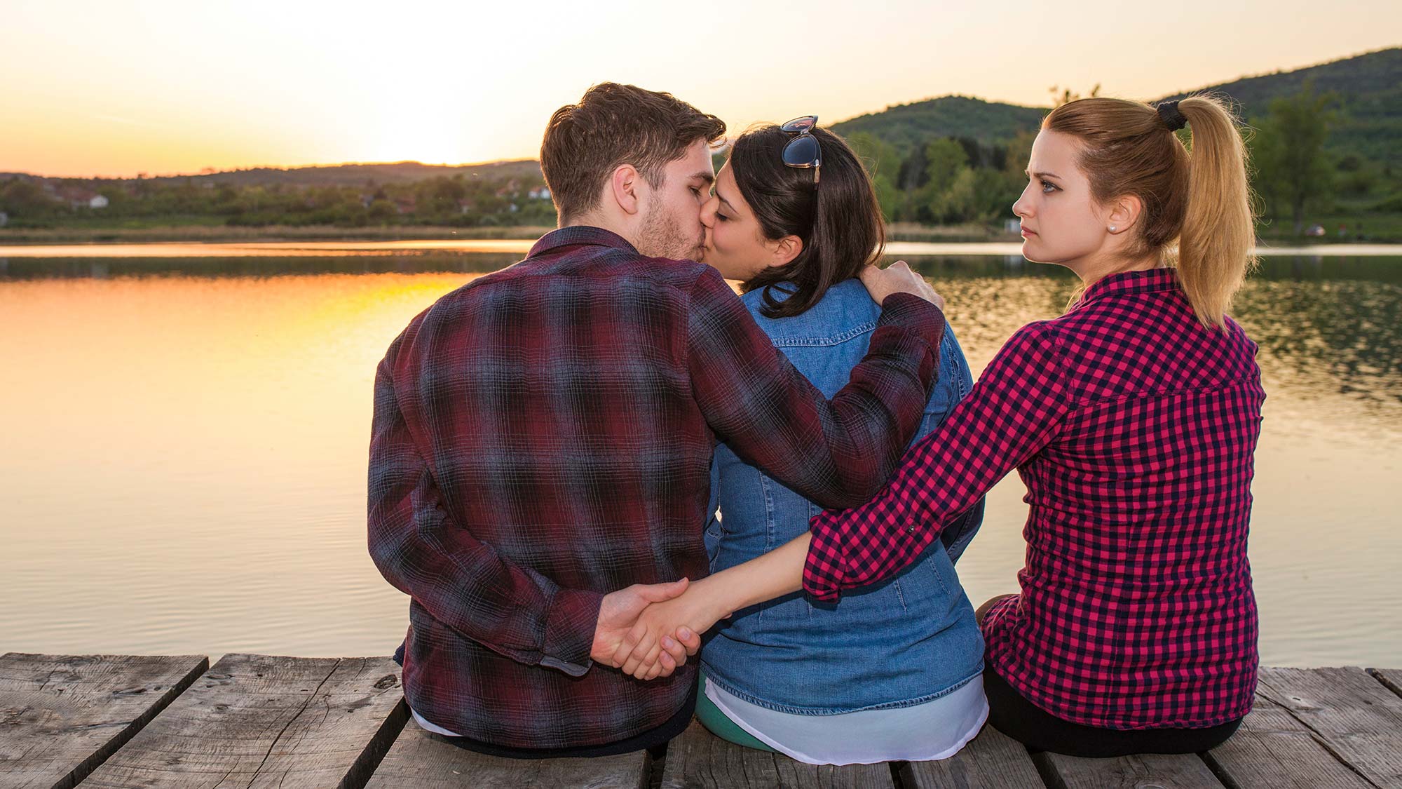 Zwei Mädchen und ein Junge sitzen auf Steg am See, die einen küssen sich die anderen beiden halten Händchen