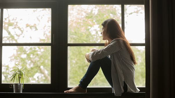 Mädchen sitzt auf Fensterbank und schaut traurig aus dem Fenster