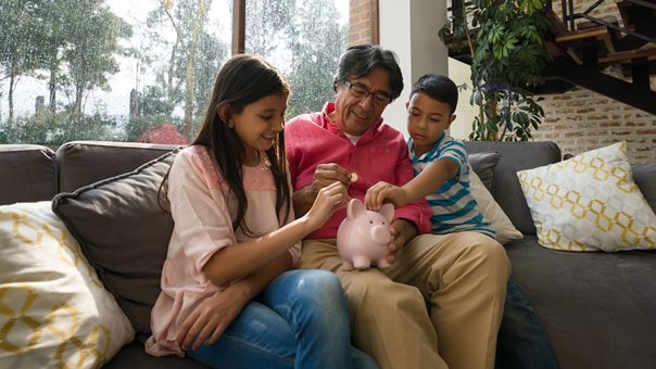 (c) iStockphoto.com/Hispanolistic Familie sitzt auf dem Sofa und schmeißt Geld in Sparschwein